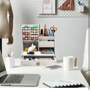 May include: White desk organizer with multiple compartments for pens, pencils, markers, and other office supplies. The organizer has a drawer and a small shelf. It is sitting on a white desk with a laptop, a notebook, and a mug.