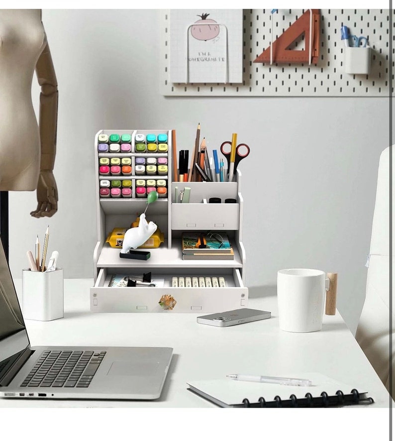 May include: White desk organizer with multiple compartments for pens, pencils, markers, and other office supplies. The organizer has a drawer and a small shelf. It is sitting on a white desk with a laptop, a notebook, and a mug.
