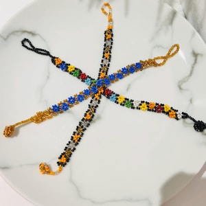 Könnte beinhalten: Sechs Armbänder mit Perlen und Blumenmustern in verschiedenen Farben, darunter Blau, Gelb, Rot und Grün. Die Armbänder sind sternförmig auf einem weißen Teller mit Marmormuster angeordnet. Jedes Armband hat an einem Ende eine Schlaufe.