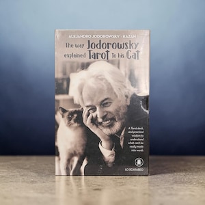 May include: A rectangular book titled The way Jodorowsky explained Tarot to his Cat by Alejandro Jodorowsky and Kazan. The cover features a black and white photograph of a man with white hair and a cat, with additional text about a Tarot deck.