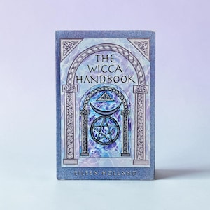 May include: A rectangular book titled The Wicca Handbook by Eileen Holland. The cover features a blue and purple marbled background with a central archway illustration containing a crescent moon, a pentagram, and a triangle symbol at the top.