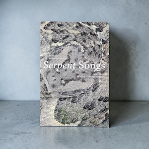 May include: A rectangular book titled Serpent Songs, an anthology of traditional craft curated by Nicholas de Matos Frisvold. The cover features a detailed, high-resolution photograph of grey, black, and white snake scales in a coiled pattern.