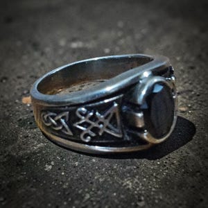 May include: A silver-toned metal ring featuring an oval black stone set in the centre. The band is decorated with engraved symbols, including a Celtic knot pattern and a geometric star-like design, with a polished finish and dark accents.