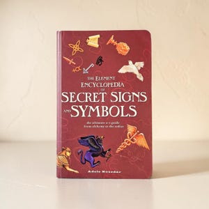 Puede incluir: Un libro de tapa dura titulado The Element Encyclopedia of Secret Signs and Symbols de Adele Nozedar. La cubierta de color rojo oscuro presenta varias ilustraciones, incluyendo una paloma blanca, un grifo morado y un caduceo.
