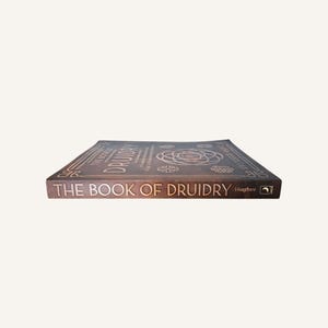 Op de afbeelding: Een bruin boek getiteld "THE BOOK OF DRUIDRY" met gouden letters. De cover heeft Keltische knoopontwerpen en de naam "Hughes". Het boek ligt plat en toont de cover en rug.