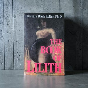 Pode incluir: Um livro intitulado "The Book of Lilith" de Barbara Black Koltuv, Ph.D. A capa apresenta uma pintura de uma mulher nua em cores escuras com o t&iacute;tulo em rosa brilhante. O livro est&aacute; em p&eacute; sobre uma superf&iacute;cie cinza.
