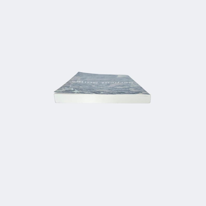 May include: A paperback book viewed from the side, showing the white pages and a cover with a dark, mottled gray and white abstract pattern. The title text Serpent Songs is printed in a light, thin font on the top cover surface.