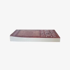 May include: A book titled "Enochian Magic in Practice" with a brown cover and white pages. The book is lying flat, with the title and author's name visible on the spine and cover. The cover features a geometric design.