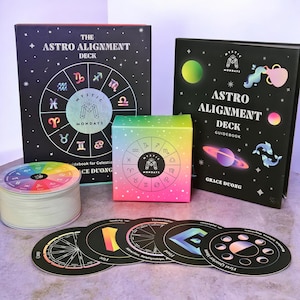 May include: A collection of astrology-themed products, including a deck of cards, a guidebook, and a box. The deck and box feature colorful designs with zodiac symbols and the text "ASTRO ALIGNMENT DECK". The guidebook is black with celestial illustrations.