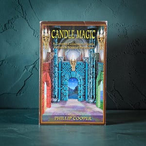 May include: A paperback book titled Candle Magic: A Coveted Collection of Spells, Rituals & Magical Paradigms by Phillip Cooper. The cover features a vibrant illustration of a stone archway with blue, red, and green pillars and golden sconces.