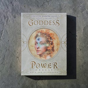 May include: A Goddess Power Oracle deck and guidebook by Colette Baron-Reid. The cover features a portrait of a woman with a third eye, surrounded by gold text and decorative elements. The deck is packaged in a light beige box.