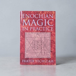May include: A book titled "Enochian Magic in Practice" with a red cover. The cover features a six-pointed star with symbols and the author's name, Frater Yechidah, at the bottom. The book is standing upright on a light gray surface.