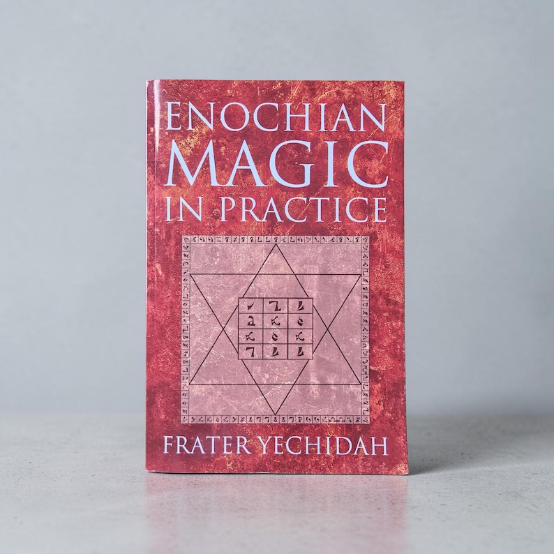 May include: A book titled "Enochian Magic in Practice" with a red cover. The cover features a six-pointed star with symbols and the author's name, Frater Yechidah, at the bottom. The book is standing upright on a light gray surface.