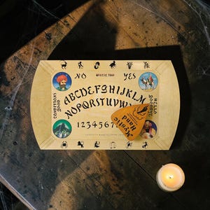 Könnte beinhalten: Ein Ouija-Brett im Vintage-Stil aus hellem Holz mit Alphabet, Zahlen und dekorativen Illustrationen. Enthält eine Planchette und die Worte "Mystic Tray", "Hello Goodbye" und "Mystic Hand". Eine brennende Kerze befindet sich in der Nähe.