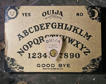 May include: A rectangular tan Ouija board featuring black lettering of the alphabet, numbers 1-9 and 0, and the words YES, NO, and GOOD BYE. A matching tan planchette with a circular center cutout rests on the board, displaying the text OUIJA.
