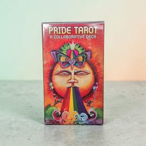 May include: A Pride Tarot deck box featuring a colorful illustration of a sun-like face with a rainbow emanating from its mouth. The box reads "Pride Tarot: A Collaborative Deck" in white text against a red background.