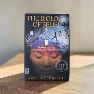 May include: A hardcover book titled "The Biology of Belief" with a dark cover featuring a graphic of a person's face and abstract figures. The book's title and author's name are displayed prominently.