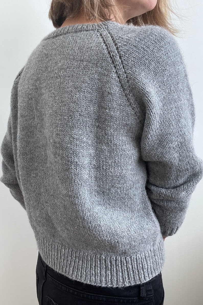 May include: A gray knitted sweater with a round neckline and long sleeves. The sweater has a ribbed hem and cuffs.