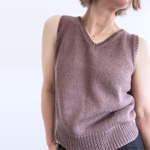 Könnte beinhalten: Eine ärmellose, mauvefarbene Strickweste mit V-Ausschnitt. Die Weste hat einen gerippten Saum und ein strukturiertes Strickmuster. Das Kleidungsstück ist aus einem weichen Material, möglicherweise Wolle. Der Stil ist lässig und bequem.
