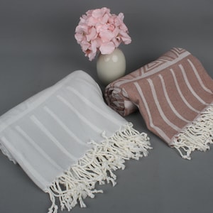 May include: Two striped towels with fringe, one white and one light brown, laid out on a gray surface. A vase with pink flowers sits behind the towels.