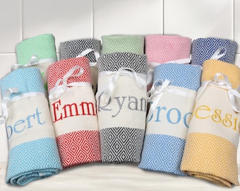 May include: Eight personalized towels rolled up and tied with white ribbon. The towels are in various colors and patterns, including blue, green, yellow, pink, and red. Each towel has a name embroidered on it, including "Bert", "Emma", "Ryan", "Brodie", and "Jessie".
