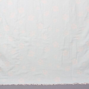 May include: A light blue beach towel with a repeating pattern of pale pink floral designs. The towel has a fringed edge along the bottom. The fabric appears soft and lightweight, suitable for use at the beach or pool.