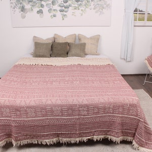 May include: A pink throw blanket with a white geometric pattern and fringe trim. The blanket is on a bed with pillows and a rug on the floor.