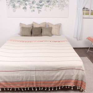 May include: A beige and white striped blanket with orange and red accents is spread across a bed. The blanket has a fringe along the bottom edge. Three pillows in shades of beige and green are on the bed.