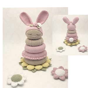 Op de afbeelding: Een handgemaakt gehaakt stapelspeelgoed in de vorm van een konijn. Het konijn is roze met lange oren en een bloem op zijn hoofd. Het speelgoed heeft verschillende stapelringen in roze en beige tinten, die op een geelgroene bloemenbasis staan. Extra gehaakte bloemen zijn aanwezig.