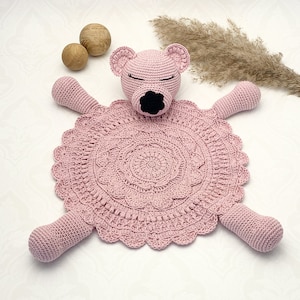 Puede incluir: Una manta de seguridad para bebé de ganchillo rosa hecha a mano con una cabeza de oso con los ojos cerrados y una nariz negra. La manta tiene un diseño circular y de encaje con cuatro brazos extendidos. Dos esferas de madera y hierba ornamental seca en el fondo.