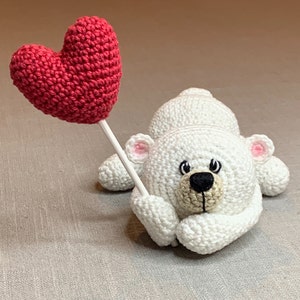 May include: A white crocheted teddy bear holding a red crocheted heart. The bear has black eyes, a brown nose, and pink ears.