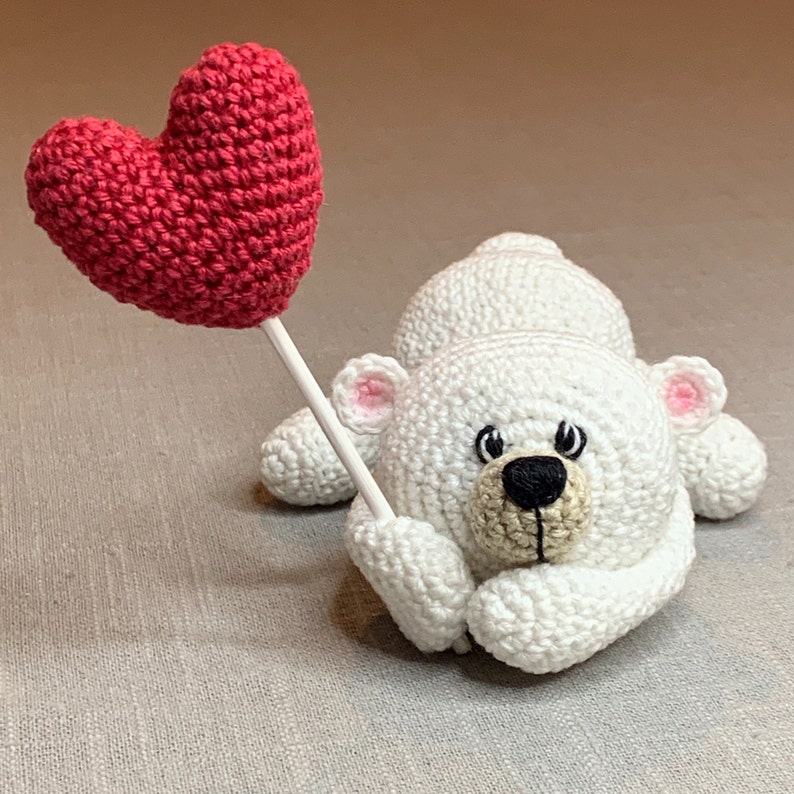 May include: A white crocheted teddy bear holding a red crocheted heart. The bear has black eyes, a brown nose, and pink ears.