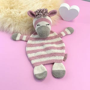 May include: A handmade crochet zebra toy in pink, white, and gray. The zebra has a striped body, a gray snout, and closed eyes. The toy is lying on a pink surface with a white heart-shaped decoration and a fluffy rug in the background.