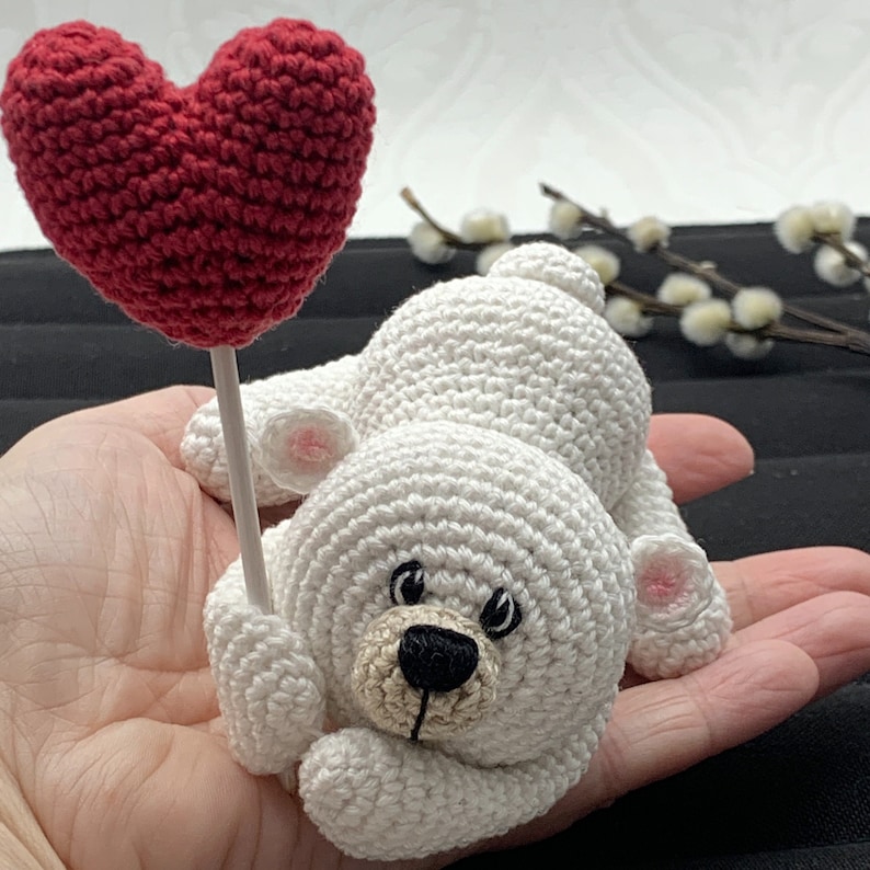 May include: A white crocheted teddy bear holding a red crocheted heart on a stick. The bear has black eyes and a brown nose.
