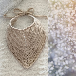 Puede incluir: Un babero bandana de ganchillo beige hecho a mano con cierre de lazo. El babero presenta un patrón texturizado en forma de cheurón y se muestra sobre una superficie blanca y suave. La estética general es sencilla y rústica.
