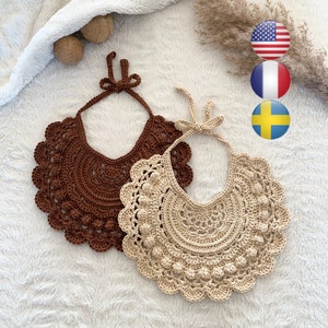 May include: Two crocheted baby bibs, one brown and one beige, with a scalloped edge and a row of small crocheted balls around the neckline. The bibs are laid out on a white fluffy surface.