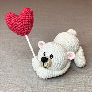 May include: A white crocheted polar bear holding a red heart-shaped crochet balloon. The bear has black eyes and a brown nose.