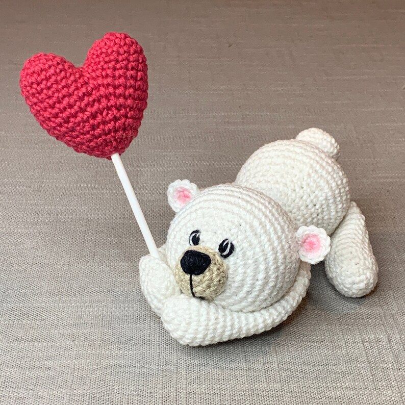 May include: A white crocheted polar bear holding a red heart-shaped crochet balloon. The bear has black eyes and a brown nose.