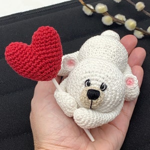 May include: A white crocheted teddy bear holding a red crocheted heart on a stick.
