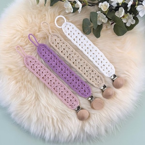 May include: Four crochet dummy clips in pastel shades: pink, purple, beige, and white. Each clip has a wooden bead and a metal clasp. The clips are displayed on a fluffy, cream-coloured surface, with white flowers in the background.