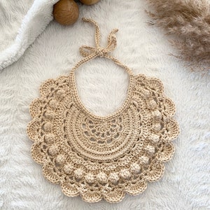 May include: A crocheted beige bib with a scalloped edge and a bow at the top.