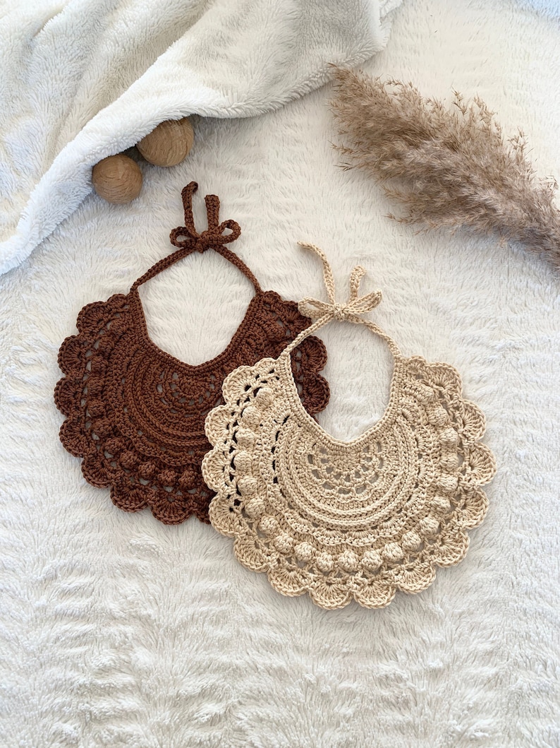 May include: Two crocheted bibs, one brown and one beige, with a scalloped edge and a bow tie. The bibs are laid out on a white fluffy surface.