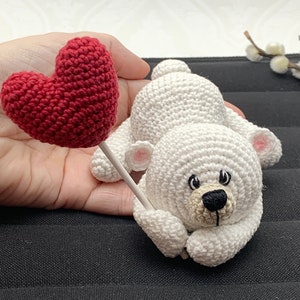 May include: A white crocheted teddy bear holding a red heart-shaped balloon. The bear has black eyes and a black nose.