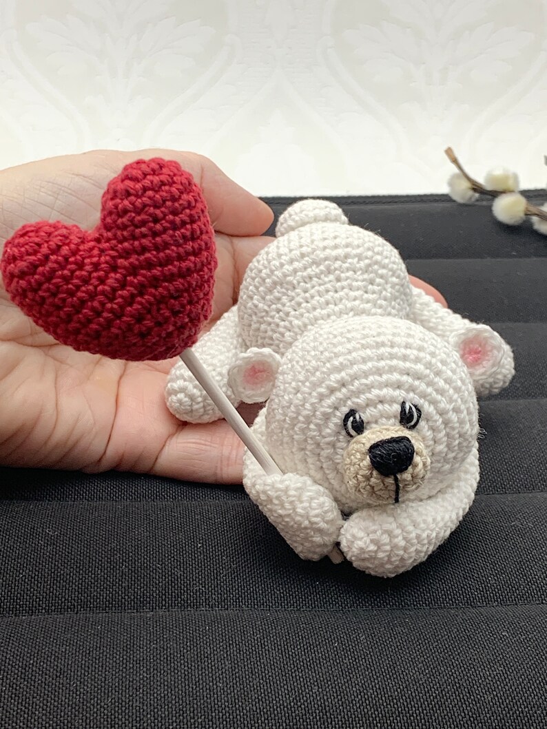 May include: A white crocheted teddy bear holding a red heart-shaped balloon. The bear has black eyes and a black nose.