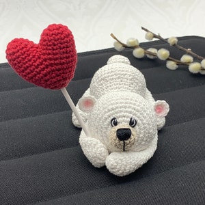 May include: A white crocheted polar bear holding a red crocheted heart. The bear is lying on a black surface.