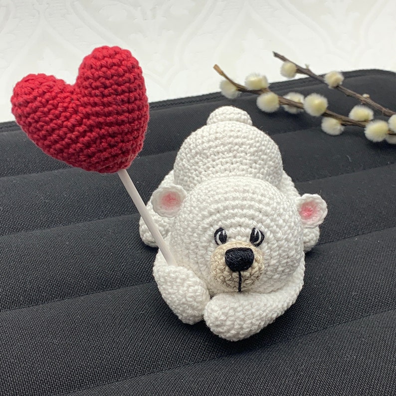 May include: A white crocheted polar bear holding a red crocheted heart. The bear is lying on a black surface.