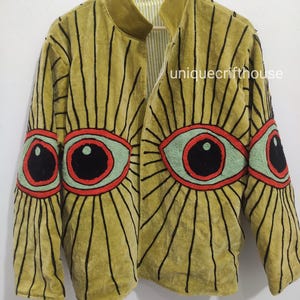 Puede incluir: Una chaqueta de terciopelo amarillo mostaza con un gran diseño de ojo bordado en la parte delantera y las mangas. Los ojos tienen iris verde claro, pupilas negras y contornos rojos con líneas negras radiantes. El texto uniquecrifthouse es visible.