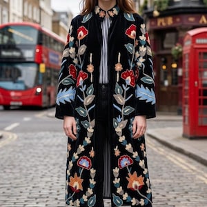 May include: A black velvet long coat featuring intricate floral embroidery in shades of red, blue, orange, and cream. The coat has a high collar, long sleeves, and a straight silhouette that falls below the knees, worn over a striped shirt and black trousers.