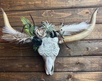 May include: A decorative steer skull with long, curved horns. The skull is painted white and adorned with a lace pattern. It is decorated with a floral arrangement of faux flowers, feathers, and greenery. The horns are a light tan color.