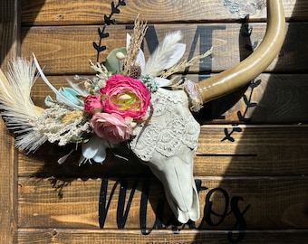 May include: A decorative faux bull skull with a floral arrangement. The skull is painted a light gray and adorned with lace. The floral arrangement includes pink and white roses, and other dried flowers. The horns are a light brown color.